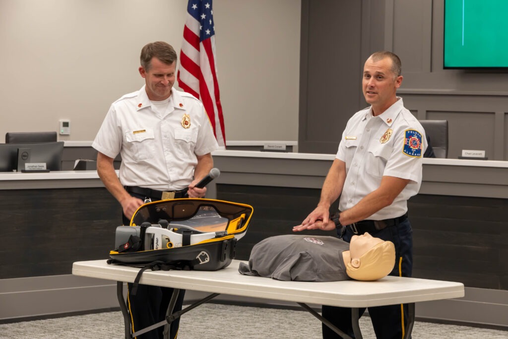 Two firefighters stand next to the LUCAS device and the CPR dummy. One mimics hand-compression on the dummy.
