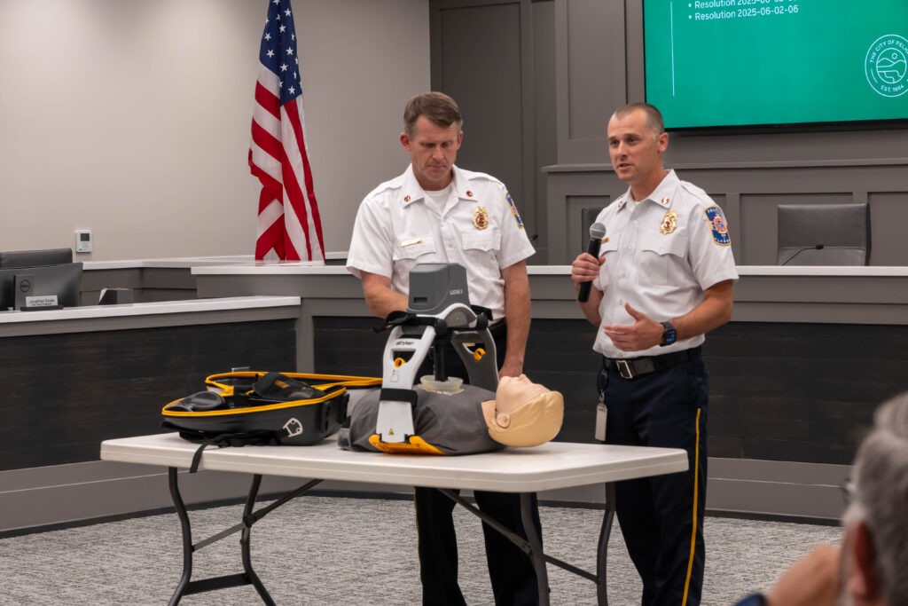 The two firefighters speak while the CPR device pushes automatic compressions on the dummy. The LUCAS device has a flat piece under the dummy with a curved section above the dummy. A compression device is connected to the curved piece and pushes on the dummy's chest.