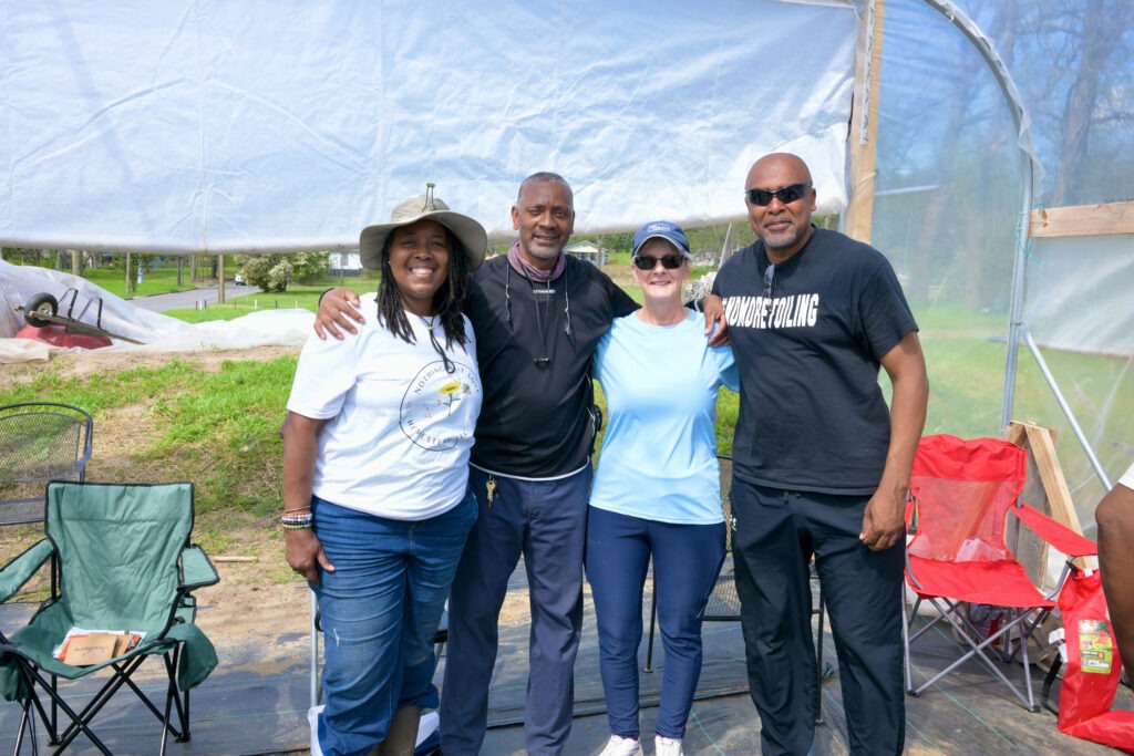 Four people pose for a picture with their arms around each others shoulders, smiling. They are standing in a covered greenhouse. There are several chairs in the background.