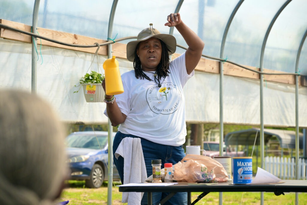 Farmer Vanessa Sanders stands in a covered greenhouse wearing a wide-brimmed hat and a t-shirt that says "Nothing But Love Homestead LLC." She is holding a funnel cake batter pitcher that she uses as a watering pitcher and standing in front of a table with old seasoning jars full of seeds and a used Maxwell House coffee tin.