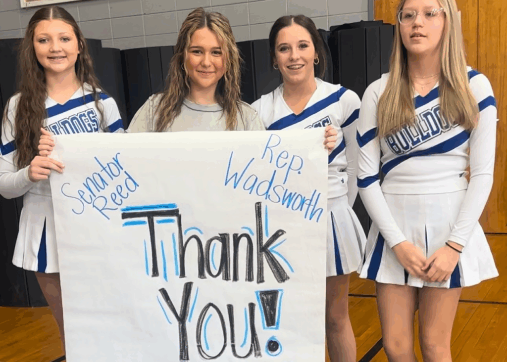 Four Jr. High cheerleaders, three of which are in uniforms, hold a paper sign designed with marker that reads "Thank You!" and the names Senator Reed and Rep. Wadsworth to the sides