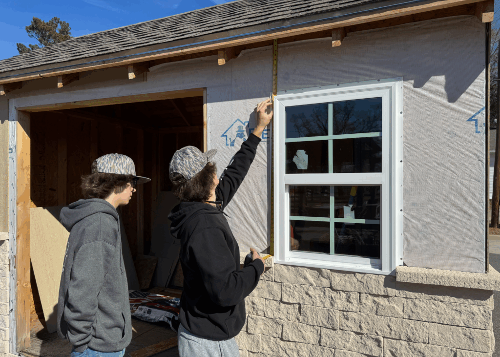 A teen boy stands in front of a garden shed, measuring the distance between already laid bricks and the roof next to a window, while another teen boy looks on.