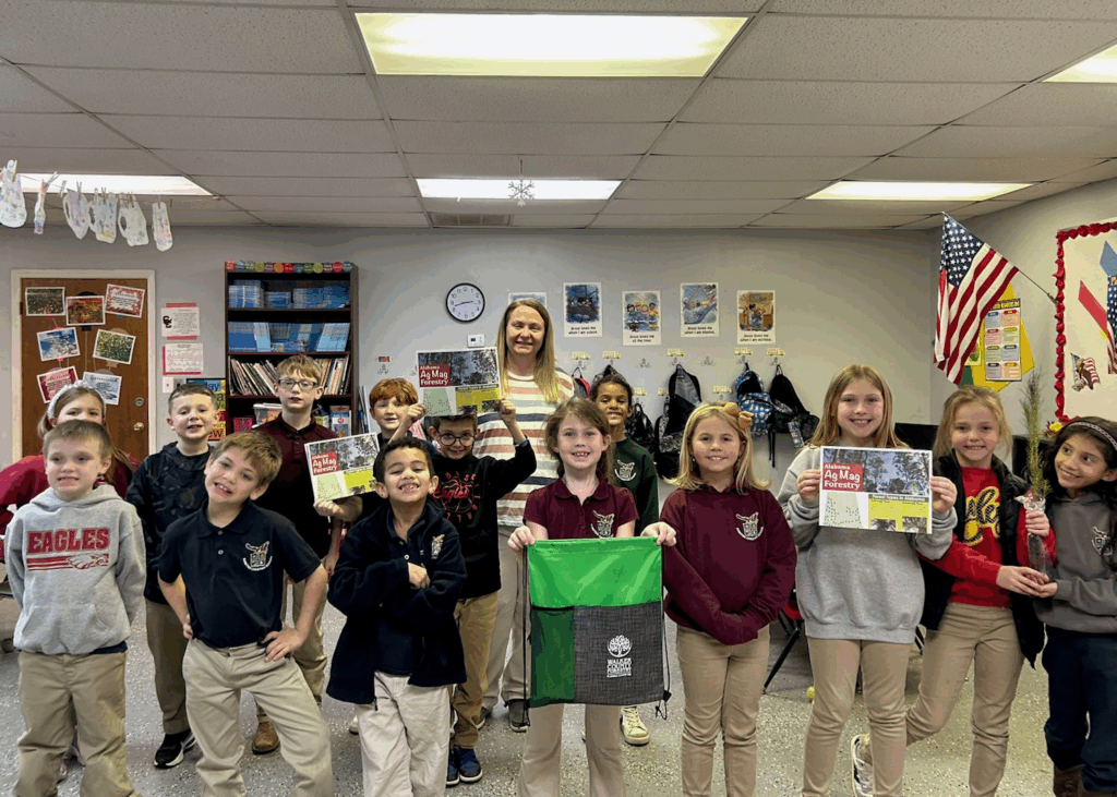 Stacy and a group of second-graders stand in a classroom smiling. Several of the students hold paper resources, one is holding a backpack, and two girls are holding a tree sapling.