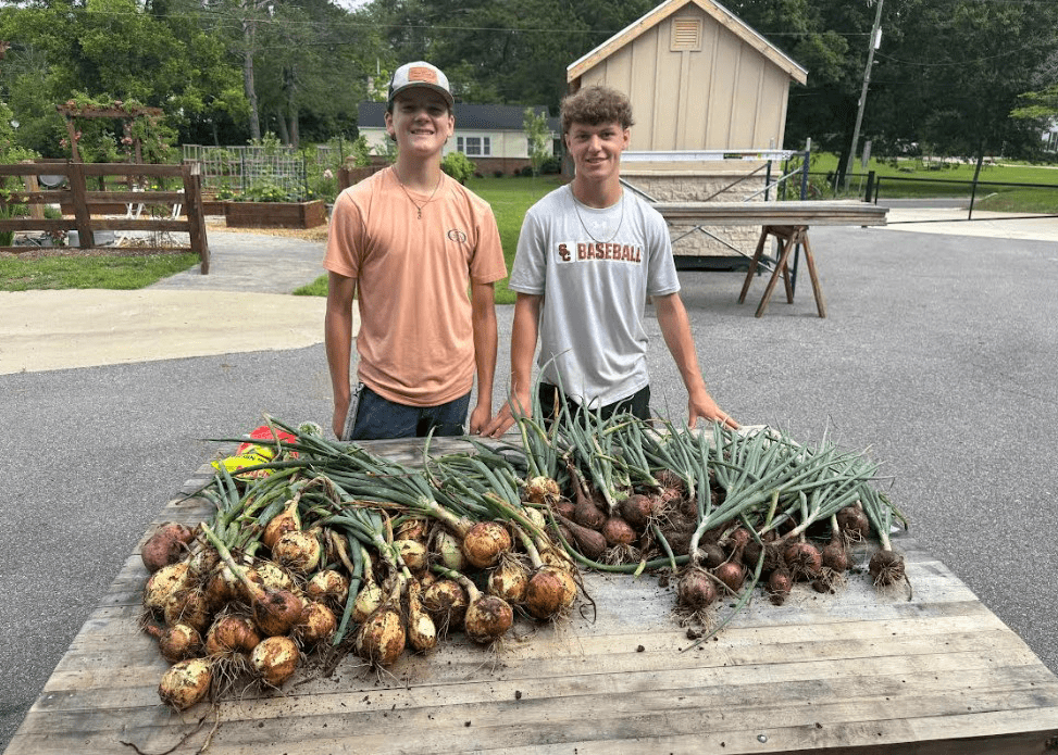 Two teen boys smile next to a table full of freshly harvested yellow and red onions. A garden shed and a flourishing fenced in community garden are visible behind them.