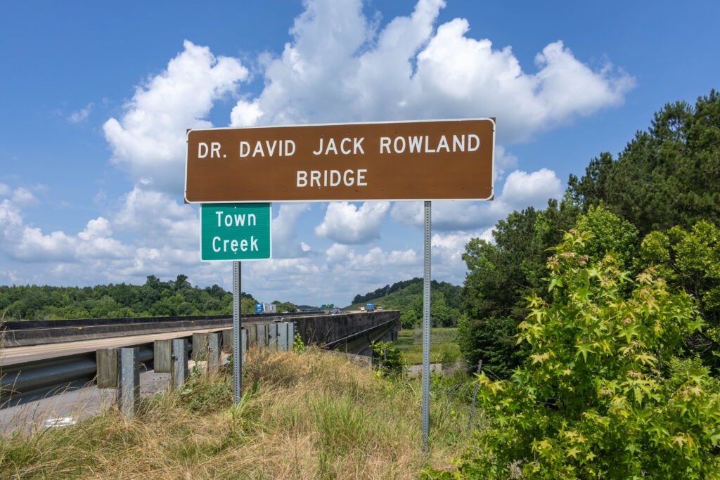 A large road sign installed next to a bridge. The main sign reads "Dr. David Jack Rowland Bridge" and a smaller sign installed underneath reads "Town Creek"