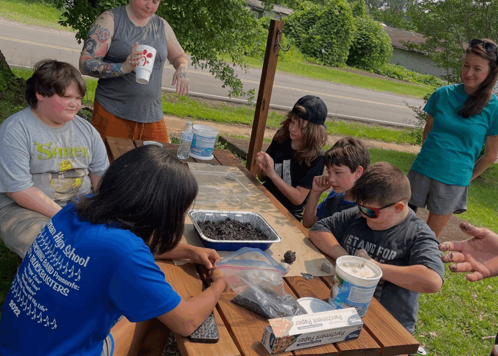 Six students sit around a picnic table that has a tin with soil, parchment paper, and several other tubs/items. A few adults look on