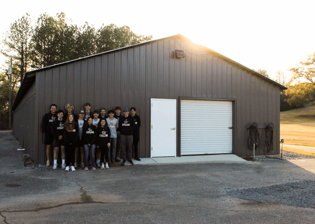 College students wearing University of Montevallo Cycling shirts stand in front of a building with metal siding. The building has a large garage door next to the regular door, as well as a bicycle cleaning station out front.