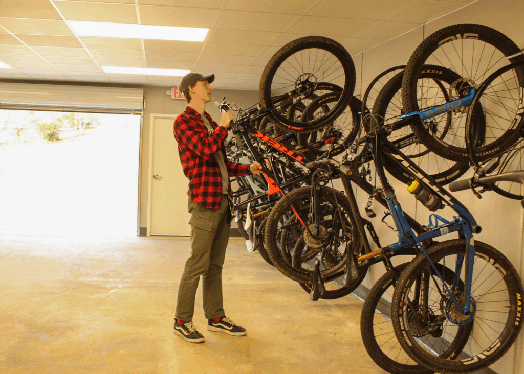 A young man stands next to a row of bikes mounted to the wall, holding onto one of the bikes as if to take it off or put it up