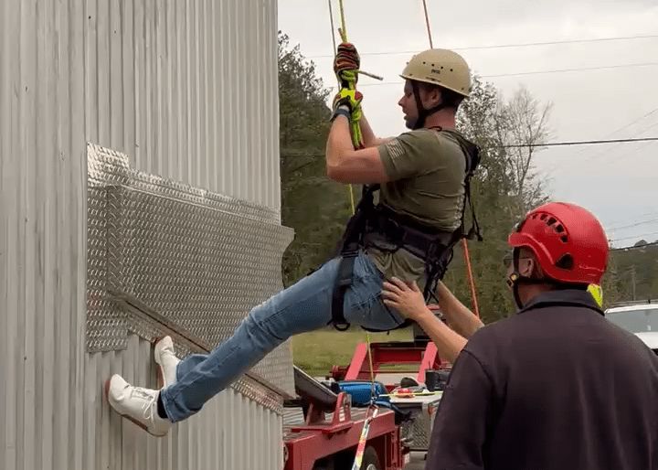 A man hangs suspended from ropes in a harness with his feet planted on an exterior wall near the bottom of a building. He is wearing a helmet, and another man in a helmet looks on while a pair of hands can be seen steadying him as he prepares to climb.
