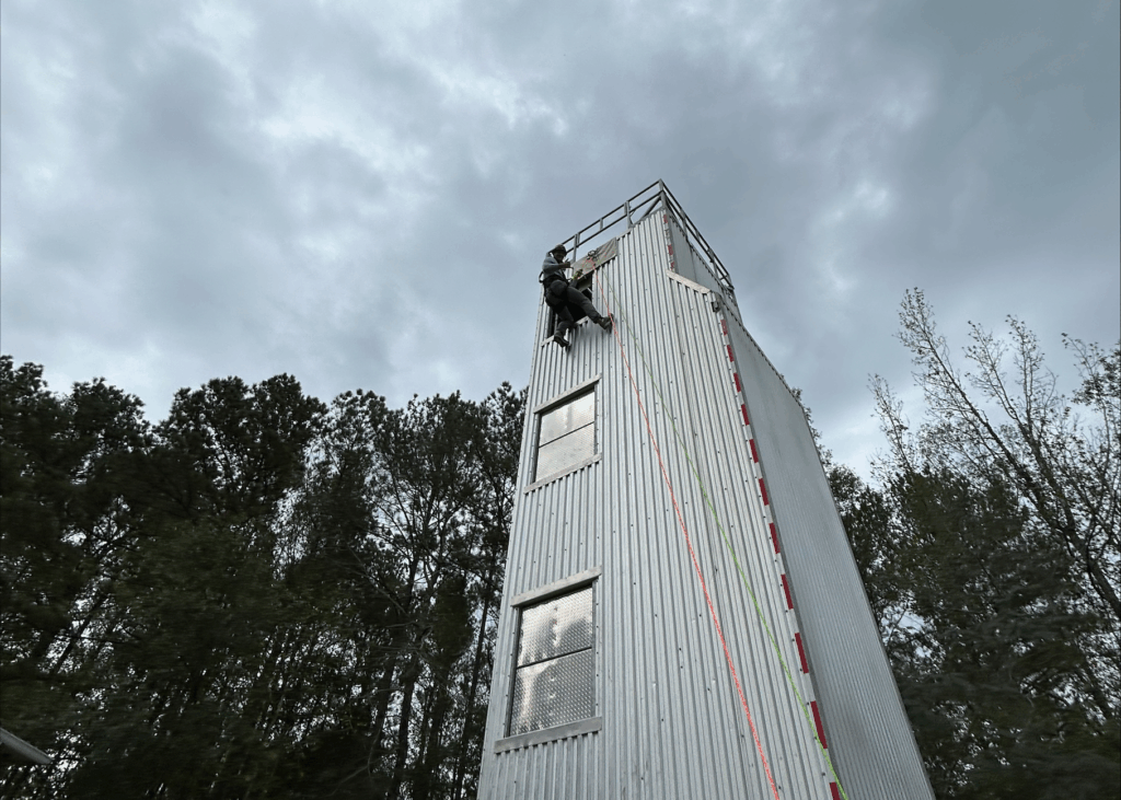 A person is suspended by ropes from the top of a tall tower building. He looks down at the camera and gives the "hang loose" hand symbol. The building is a similar height to the trees in the background