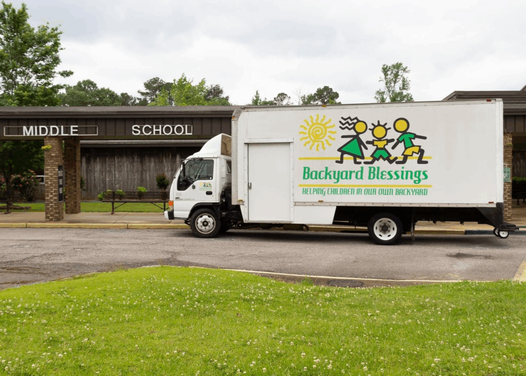 A large storage truck parked in front of Sumitan Middle School. The trucck has a large image of three stick figure children and a swirly sun above the words "Backyard Blessings" "Helping children in our own backyard"