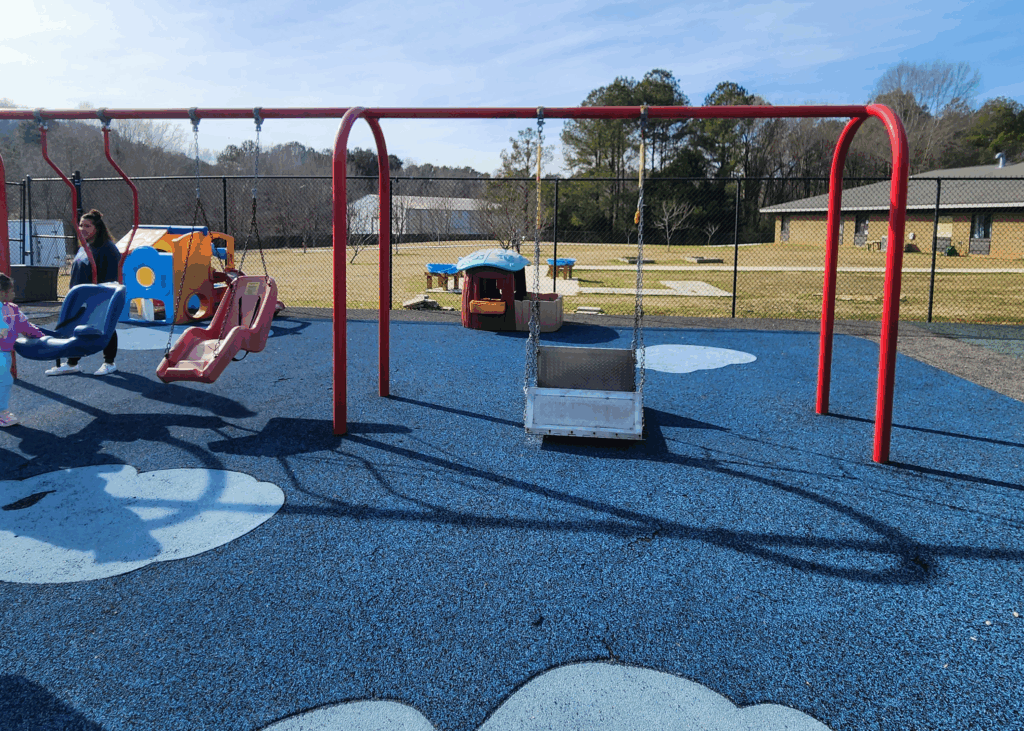 A fenced in playground with a surface made of blue pour-in-place rubber that is peeling and has chunks missing  next to playground equipment swings