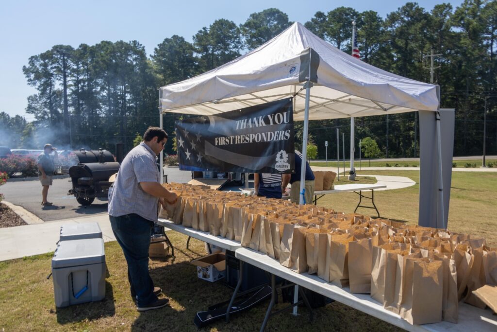 Volunteer preparing rows of brown paper lunch bags on tables under white tent with 'Thank You First Responders' banner, with grills smoking in background at outdoor event