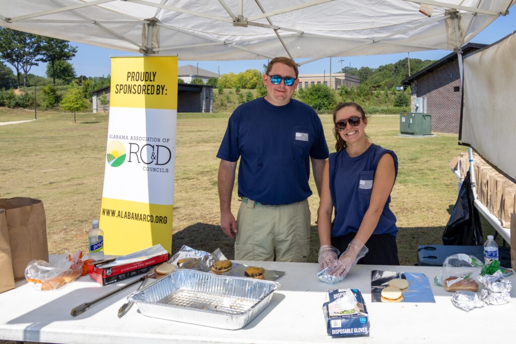 Two volunteers in matching navy shirts with disposable gloves preparing food at serving table under white tent, with Alabama Association of RC&D Councils banner displayed beside them