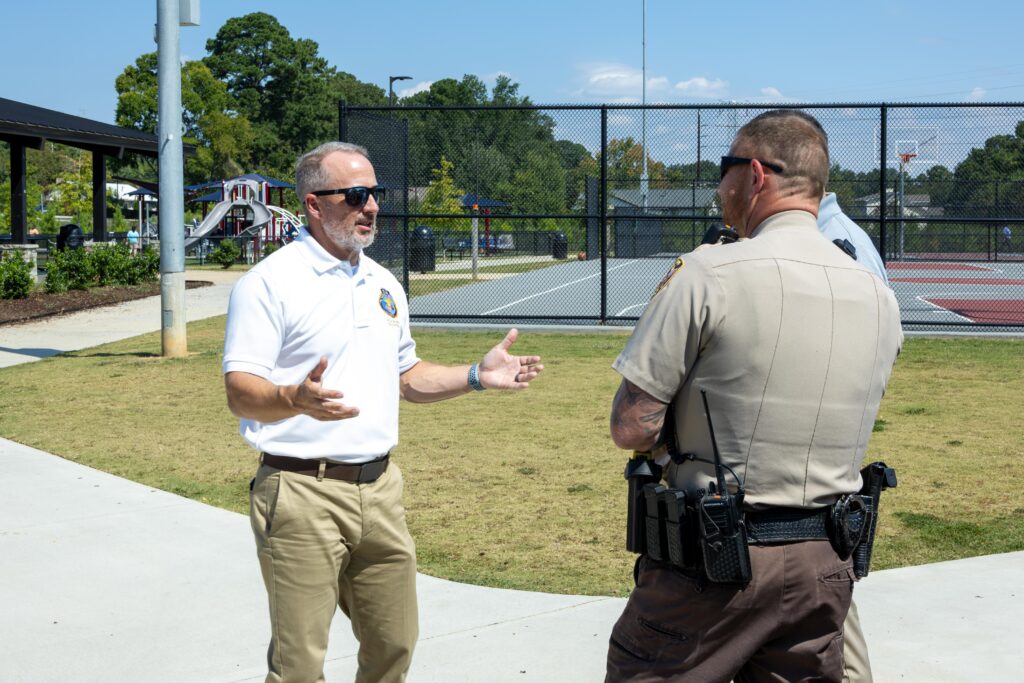 Rep. Russell Bedsole speaks with two uniformed officers. A shaded pavilion, a playground, and tennis courts are visible in the background