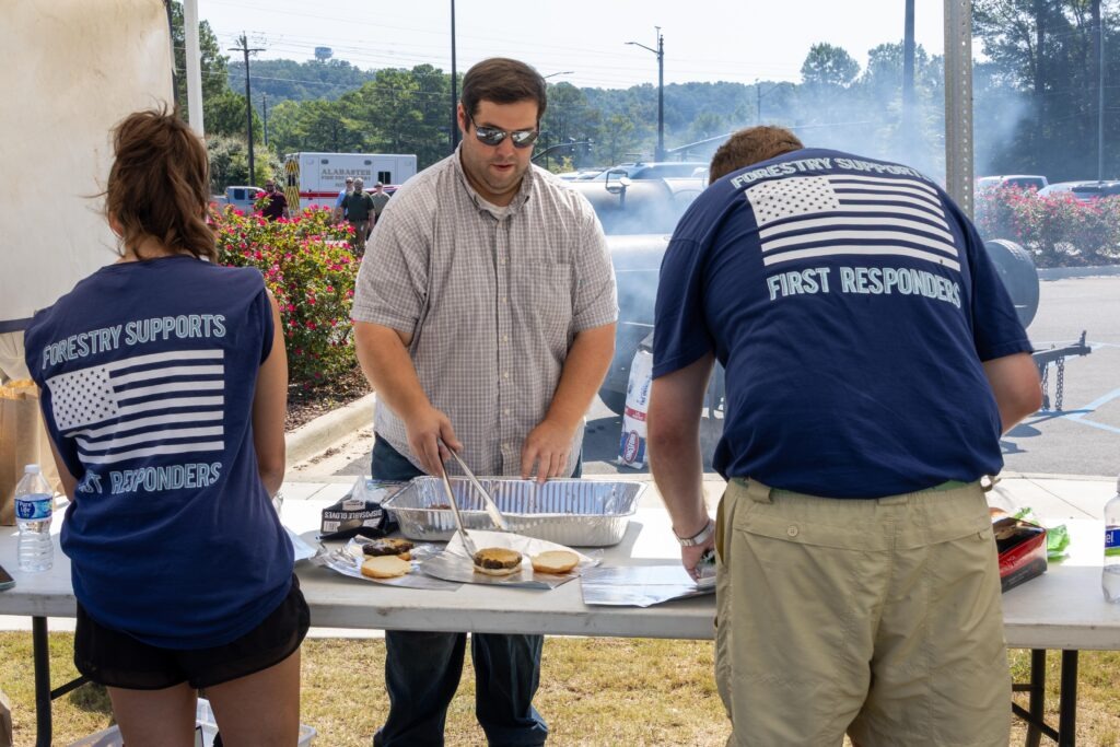 Alabama Forestry Association volunteers in matching navy 'Forestry Supports First Responders' shirts grilling hamburgers at outdoor serving station with smoke visible in background
