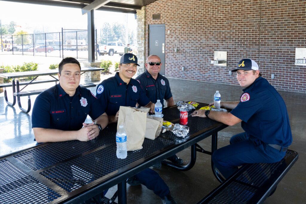 our firefighters in navy uniform shirts enjoying lunch at covered pavilion table with brown bag meals, water bottles, and soft drinks
