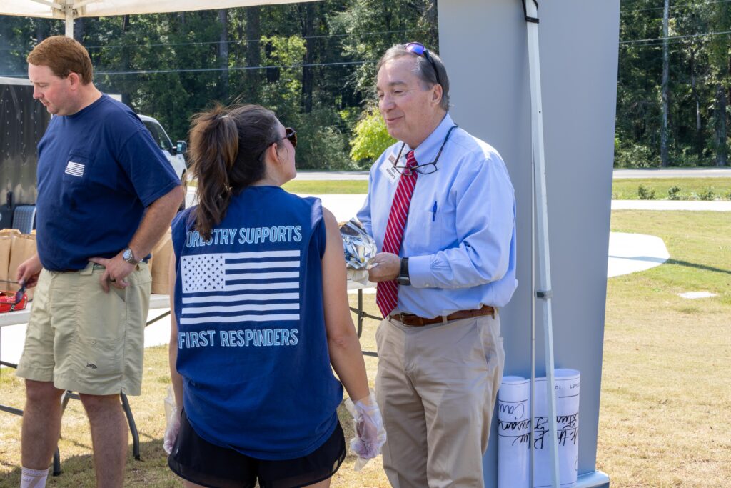 Sen. Dan Roberts in dress shirt and red tie speaking with volunteer in 'Forestry Supports First Responders' shirt at outdoor pavilion during cookout event