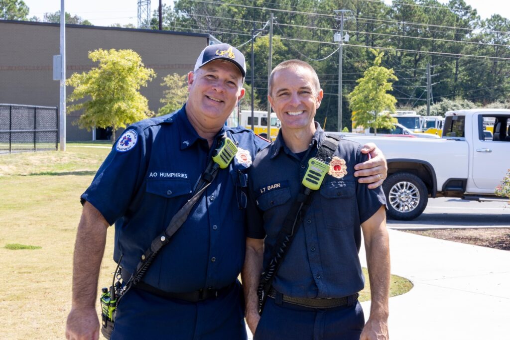 Two uniformed emergency responders with radios and equipment posing together outdoors with arms around each other's shoulders, smiling at camera