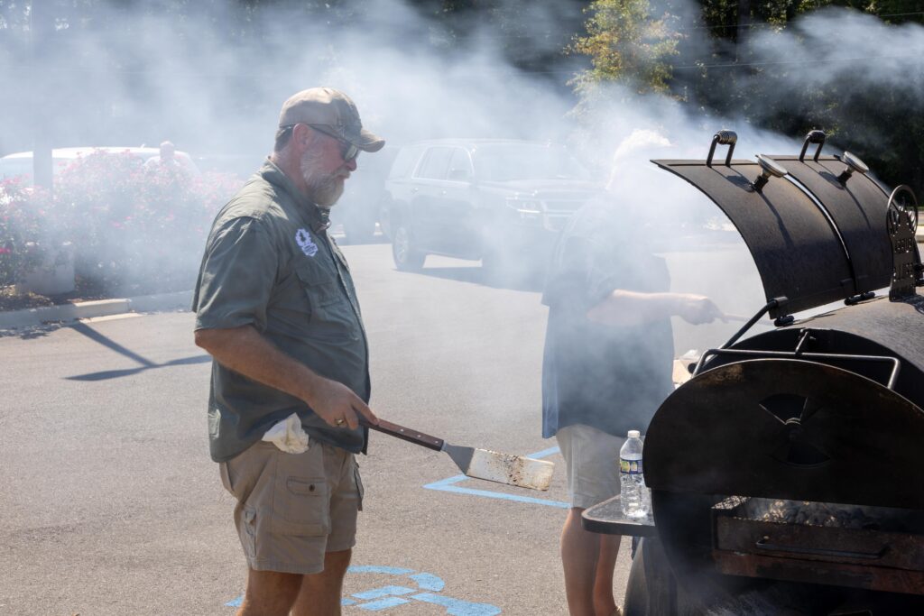 Volunteer in cap and sunglasses tending to large black smoker grill with thick white smoke billowing out during outdoor cookout event