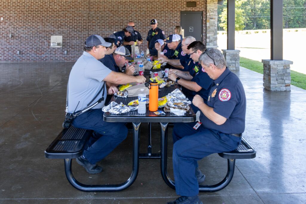 First responders in uniform sitting around picnic tables under a covered pavilion, eating lunch with aluminum foil-wrapped food, drinks, and condiments spread across the tables.