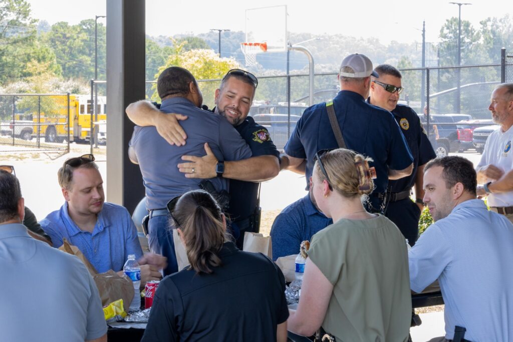 First responders and community members socializing at outdoor pavilion during appreciation cookout, with two uniformed officers embracing in foreground while others sit at picnic tables eating lunch