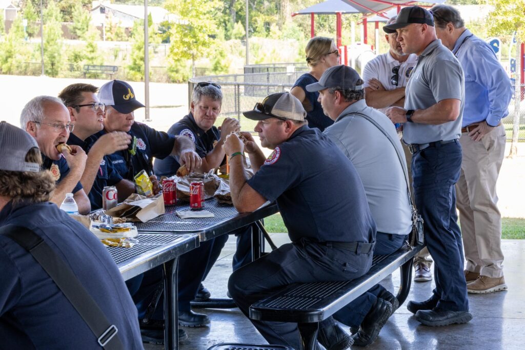 Group of first responders and community members socializing around picnic tables under a pavilion, with some standing and others seated, sharing a meal together.
