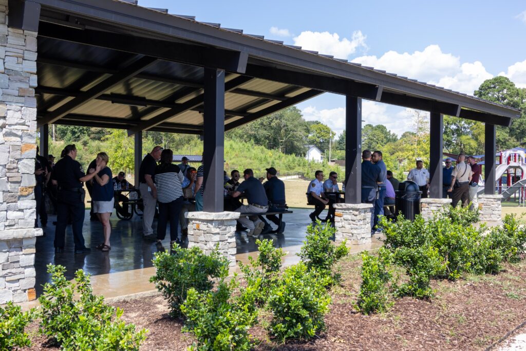 Wide view of a stone and wood pavilion filled with first responders and community members gathering for an outdoor meal, with a playground visible in the background.