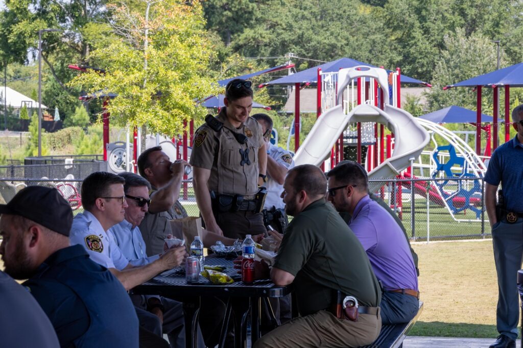 Sheriff's deputy standing and talking with seated first responders at a picnic table, with a colorful playground structure visible behind them in the park setting