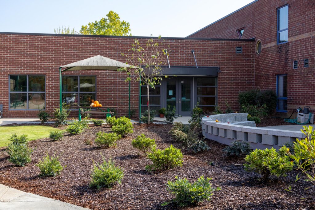 Renovated courtyard at Trace Crossings Elementary School showing landscaped garden beds with native plants, concrete block seating areas, green shade structures, and clean walkways replacing previously overgrown space.