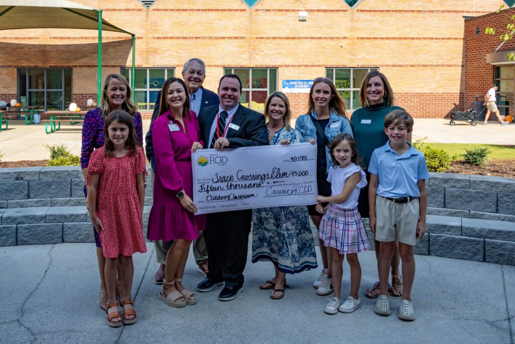 Officials and families posing with large ceremonial check from Cawaco RC&D made out to Trace Crossings Elementary for $15,000 for outdoor classroom project, with students and adults smiling in front of school building.