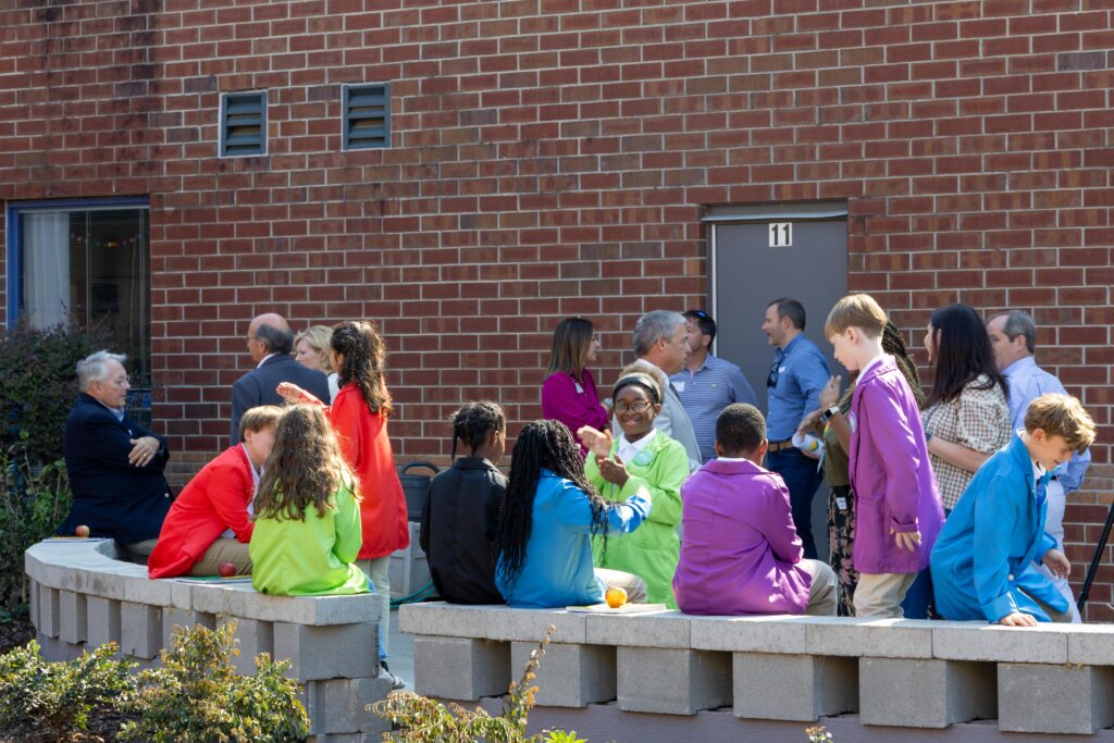 Students in colorful jackets sitting on new concrete block benches in the outdoor classroom space, with adults mingling in background during the ribbon cutting celebration.