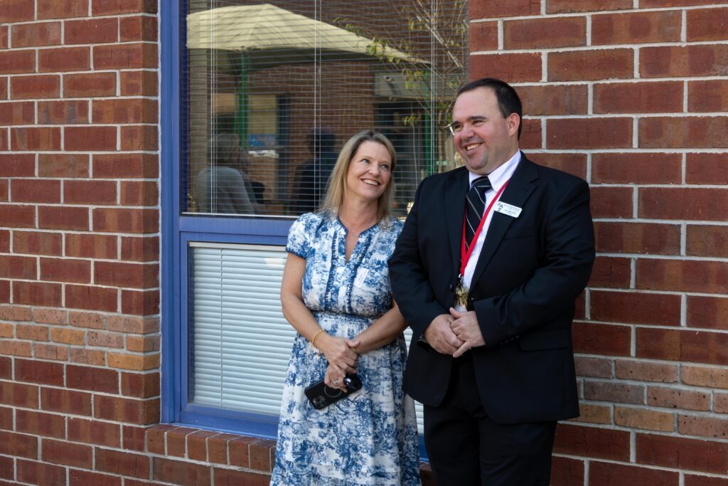 Rep. Leigh Hulsey and Principal Carl Berryhill standing together and smiling in front of brick school building during the ribbon cutting event.