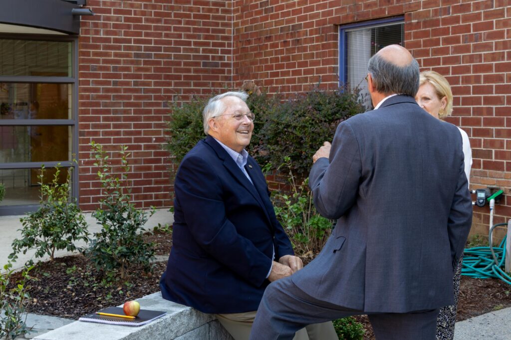 Rep. Arnold Mooney conversing with other attendees while seated on the new concrete block benches in the renovated courtyard, demonstrating the functional seating areas for outdoor learning.