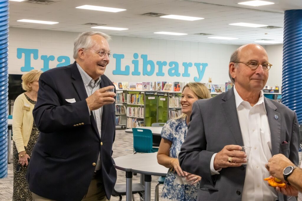 Interior view of school library with officials touring the facility, showing bookshelves, reading areas, and "Trace Library" signage on the wall as part of the reception after the ribbon cutting