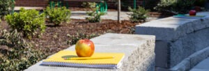 Close-up of school supplies including a red apple, yellow notebook, and pencil placed on concrete bench surface, with landscaped garden areas and shade structures visible in background, symbolizing the outdoor learning environment.