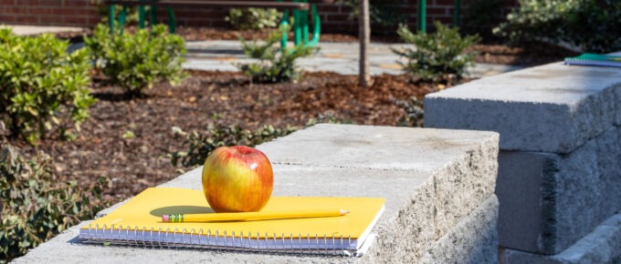 Close-up of school supplies including a red apple, yellow notebook, and pencil placed on concrete bench surface, with landscaped garden areas and shade structures visible in background, symbolizing the outdoor learning environment.