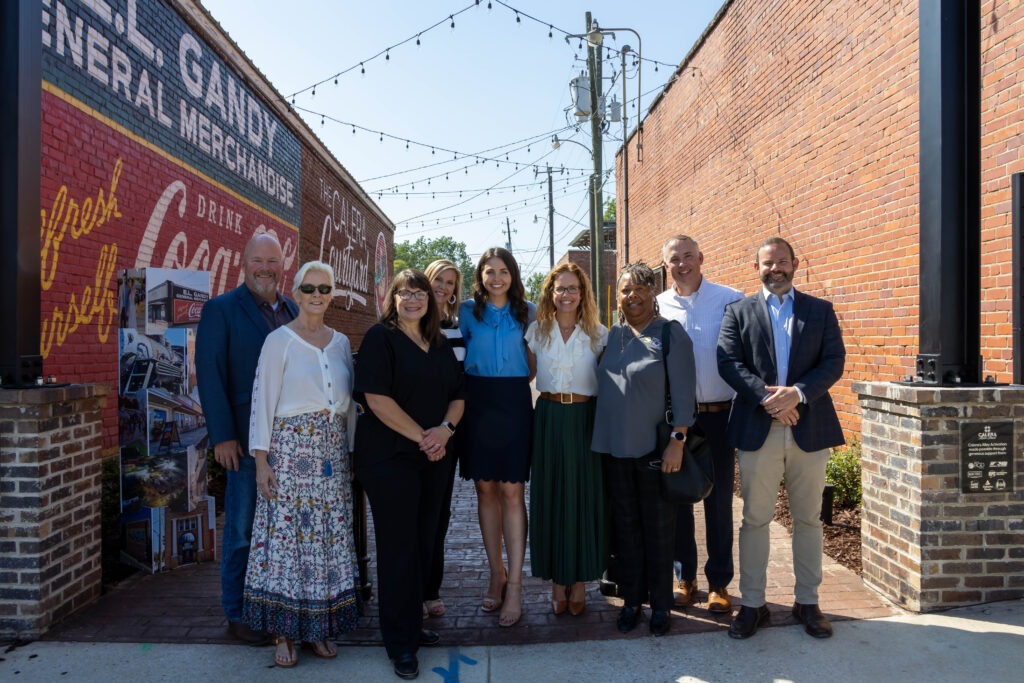 Group of eight people posing together in an alleyway with brick walls, including a vintage Coca-Cola mural, and string lights overhead