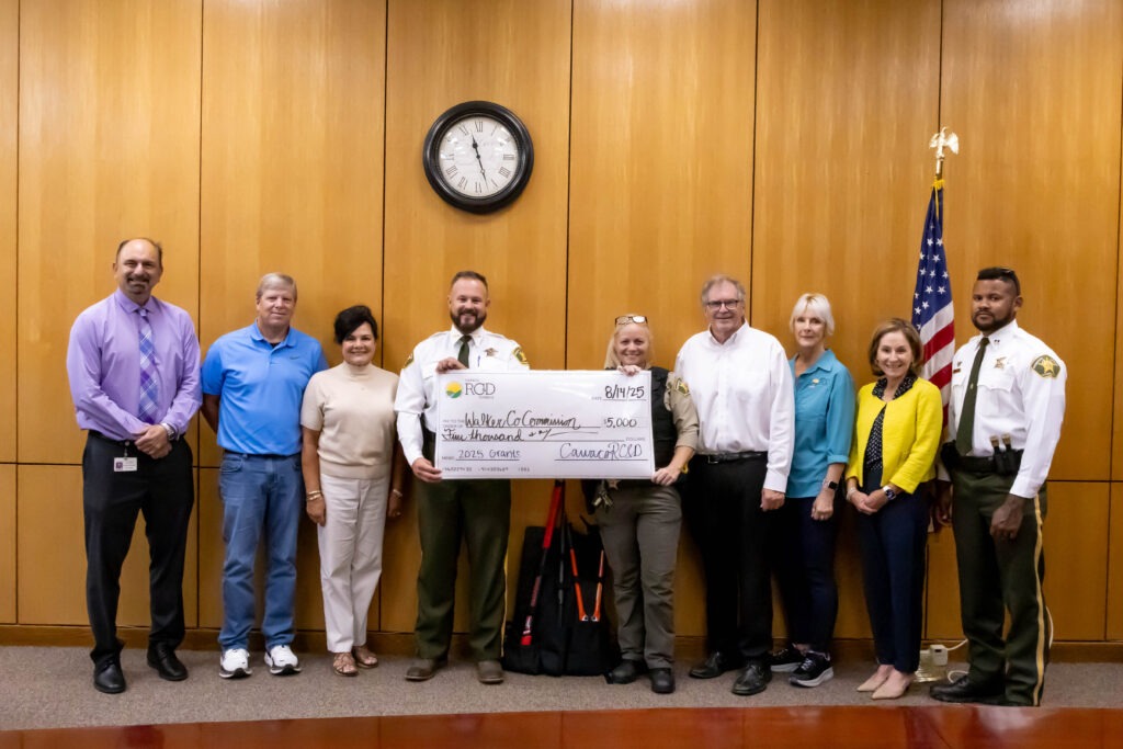 A group of nine people including school resource officers, county officials, and community representatives pose with a ceremonial check for $5,000 from Cawaco RC&D to the Walker County Commission, dated August 14, 2025, with an American flag visible in the background.