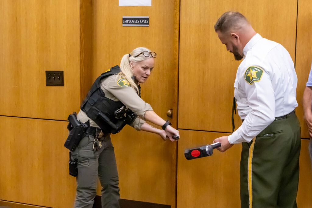 Two law enforcement officers demonstrate proper handling of breaching tools with black and red handles near an "Employees Only" door in a wood-paneled hallway.