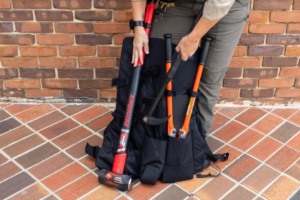 Close-up view of a tactical backpack on brick pavement displaying organized breaching tools including a red sledgehammer and orange-handled pry bars in specialized pouches, with hands demonstrating the storage system.