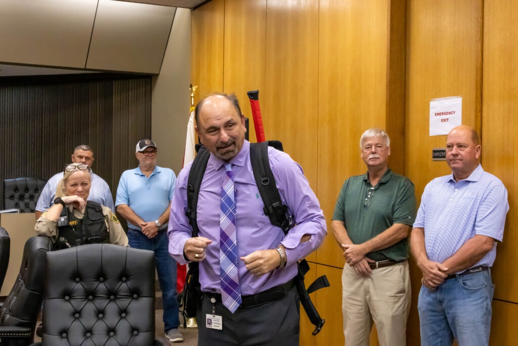Superintendant Willingham in a purple dress shirt and plaid tie speaks while wearing the tactical backpack