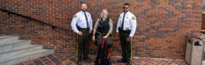 Three Walker County Sheriff's Office personnel in uniform pose outside the Walker County Courthouse brick entrance, with a deputy in the center holding an red-handled breaching tool from a tactical backpack kit.