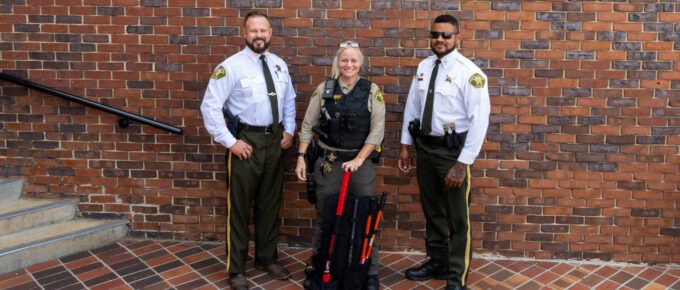 Three Walker County Sheriff's Office personnel in uniform pose outside the Walker County Courthouse brick entrance, with a deputy in the center holding an red-handled breaching tool from a tactical backpack kit.