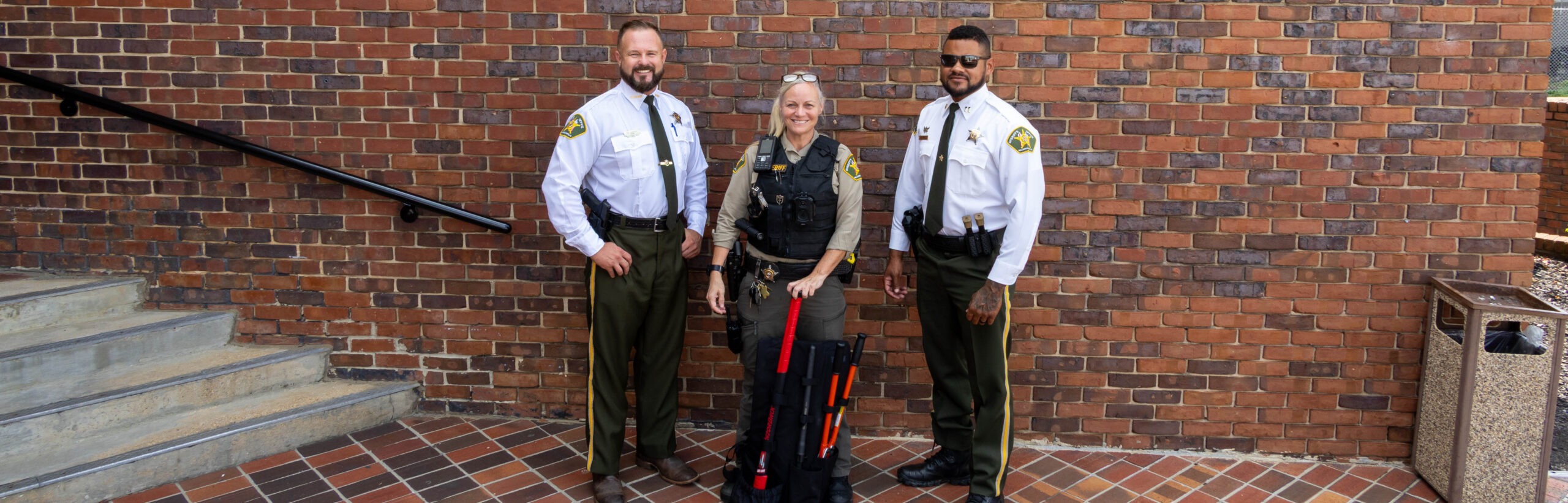 Three Walker County Sheriff's Office personnel in uniform pose outside the Walker County Courthouse brick entrance, with a deputy in the center holding an red-handled breaching tool from a tactical backpack kit.