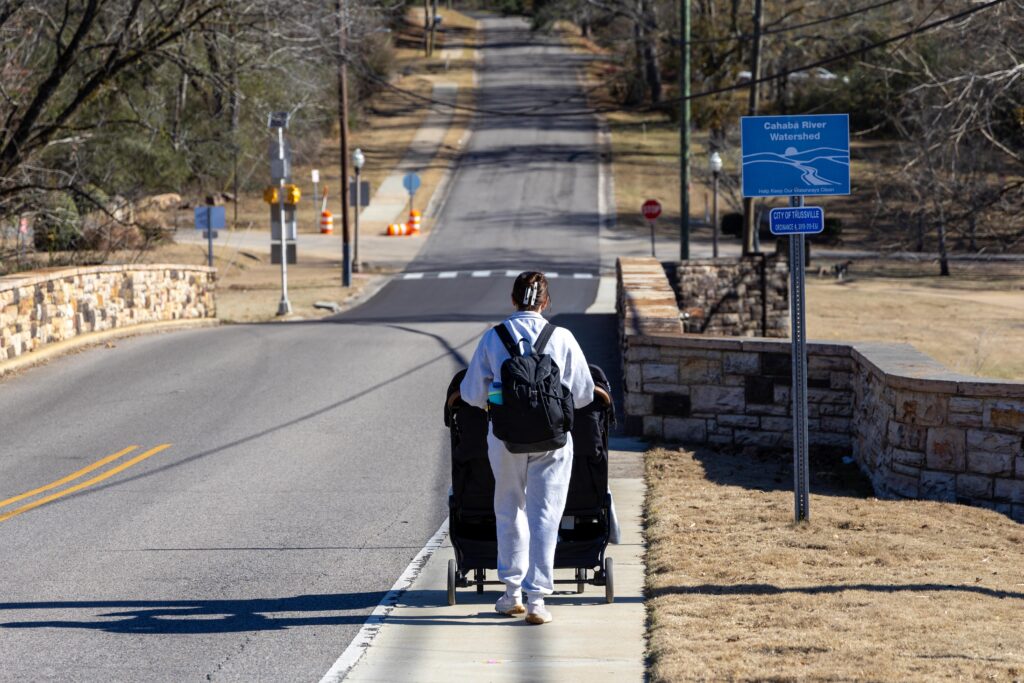 A mother walks away from the camera, pushing a two-seater stroller down a sidewalk towards a historic bridge. A sign next to the sidewalk reads Cahaba River Watershed"