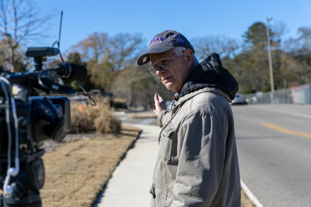 Man in windbreaker and a hat stands sideways, gesturing behind him to the sidewalk he is standing on. 