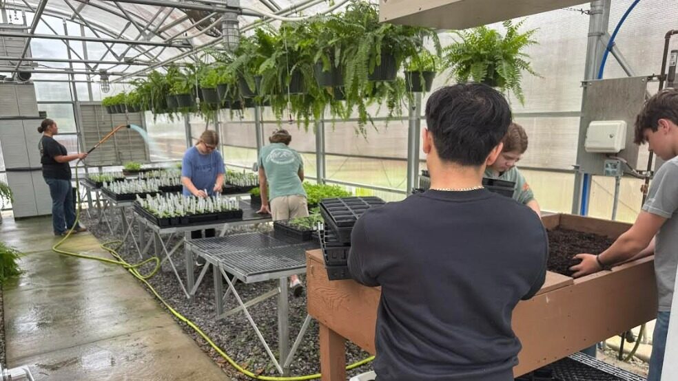 Six interns work in a greenhouse doing various tasks, including watering, planting, and labeling plants