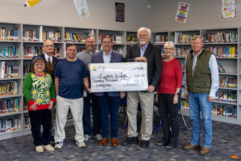 Group of eight people stand in front of newly shelved library bookcases, holding a ceremonial check for $20,000 from Cawaco RC&D made out to Lupton Jr. High for a Library Renovation, dated 3/16/26.
