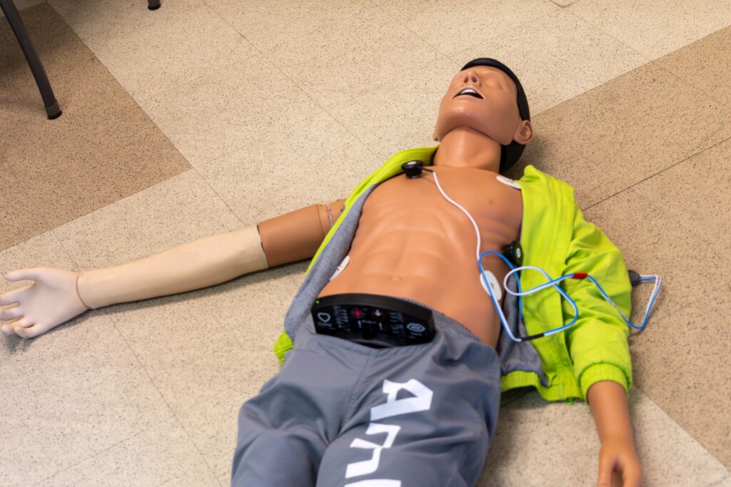 An AmbuMan Advanced CPR and emergency medical training manikin lies on a tile floor with its chest exposed. The manikin wears gray shorts and a partially open yellow-green jacket. Medical training electrodes and wires are attached to the chest, and an electronic monitoring device is clipped to the waistband.